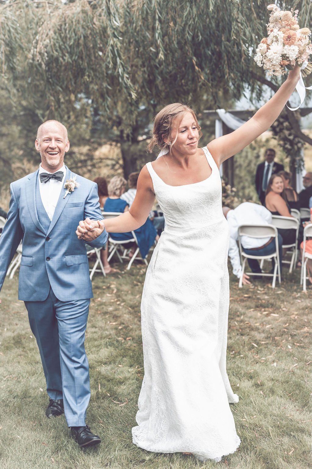 A couple walking down the aisle after being married and the bride is joyfully raising her bridal bouquet with her new husband in tow.